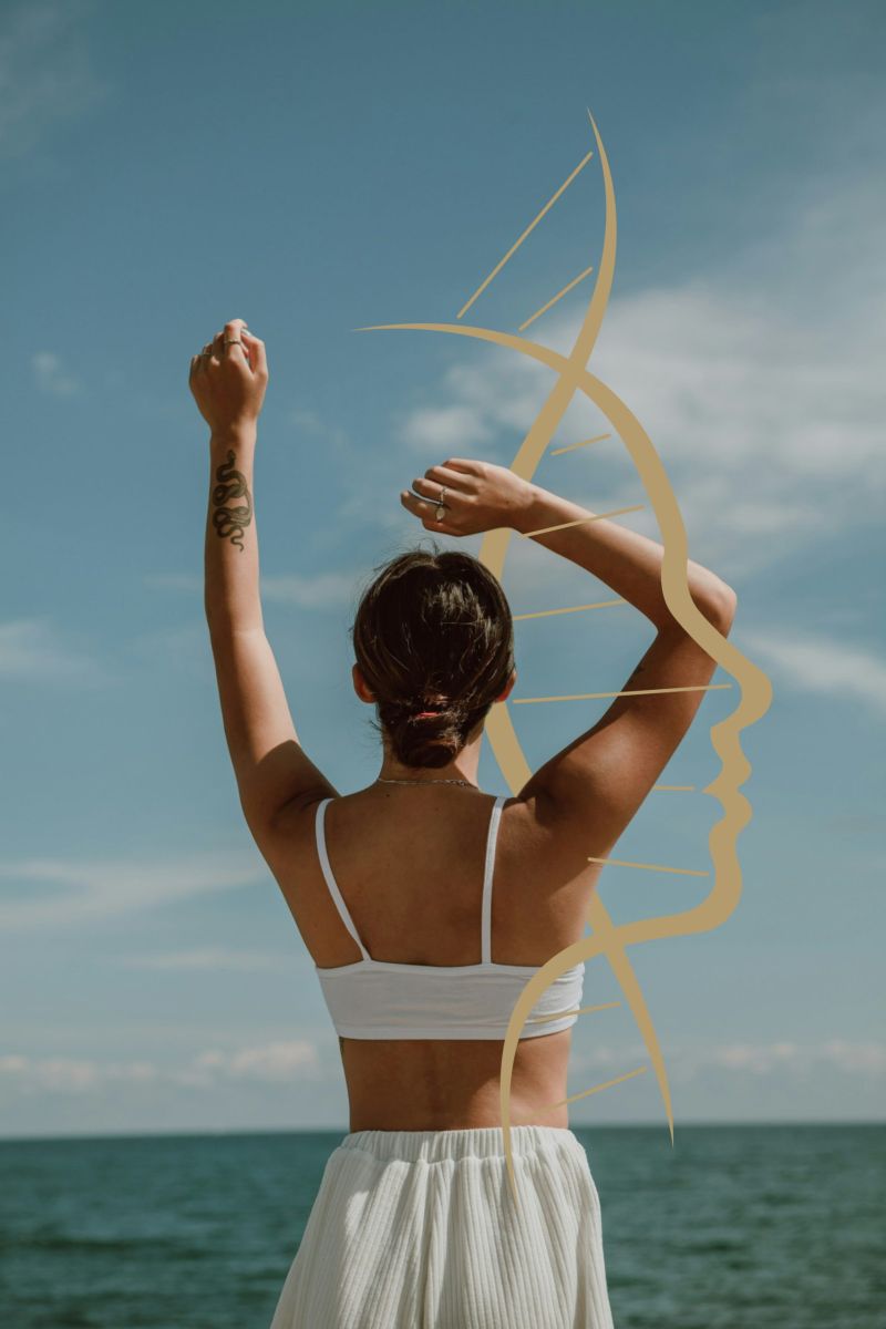 Woman in a white top stands on the beach, arms raised joyfully against a clear blue sky.