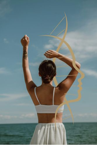 Woman in a white top stands on the beach, arms raised joyfully against a clear blue sky.
